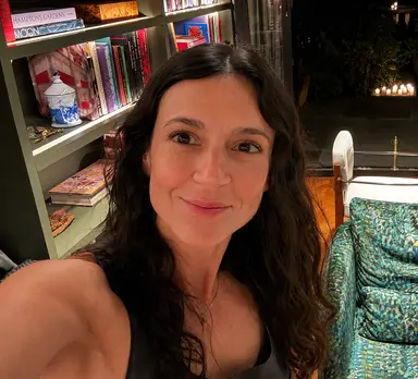 Smiling woman with long dark wavy hair in front of a bookshelf and patterned armchair.