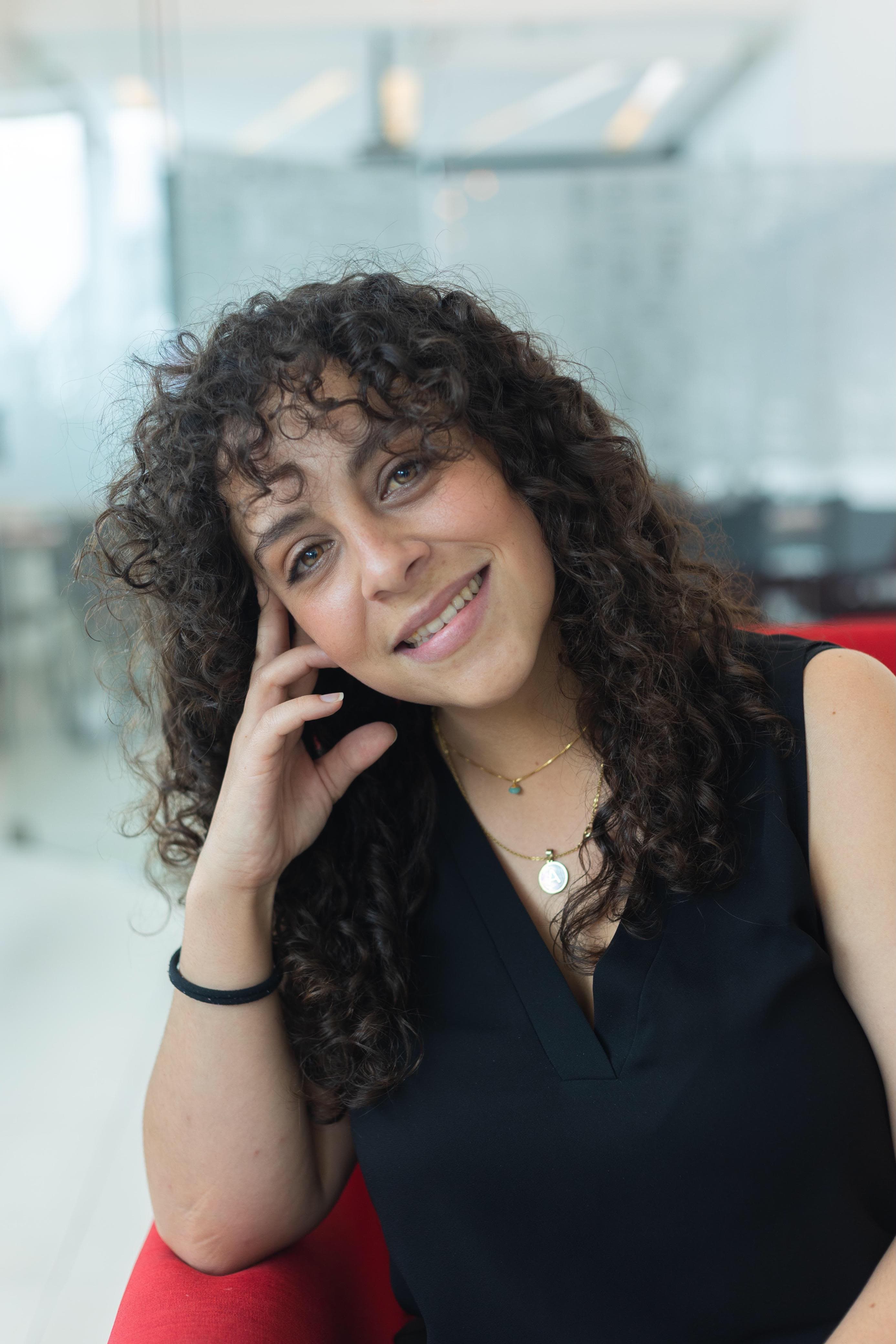 Smiling woman with dark curly hair and gold necklaces resting her hand on her face.