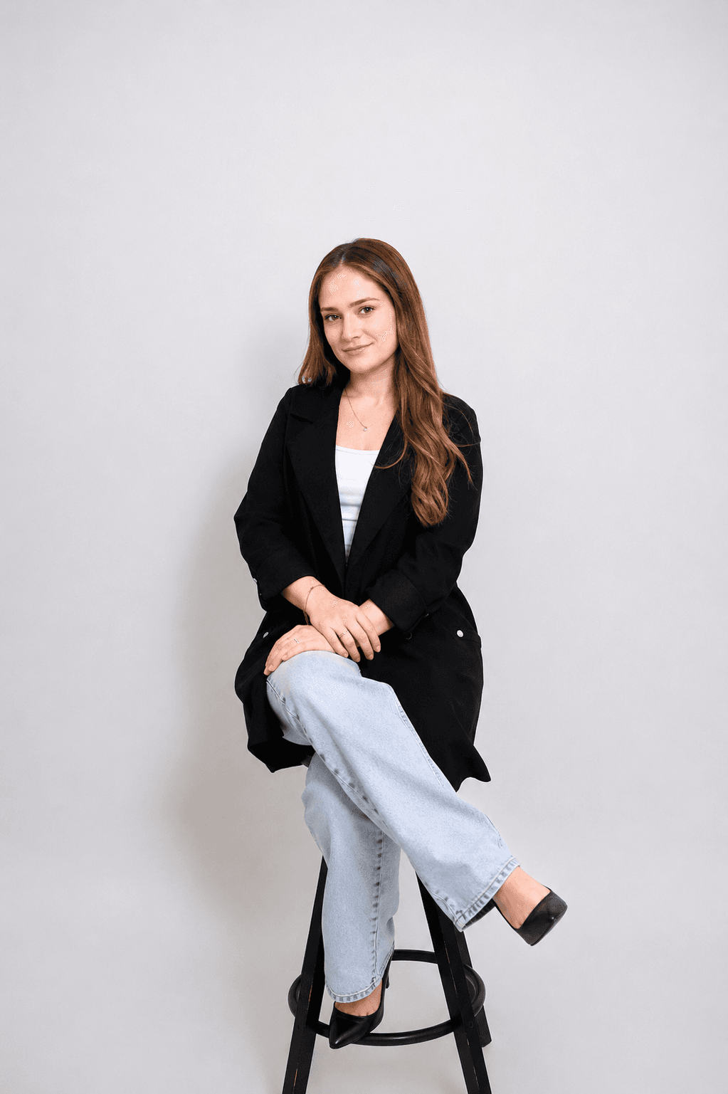 Woman with long brown hair in a black blazer and jeans sitting on a stool.