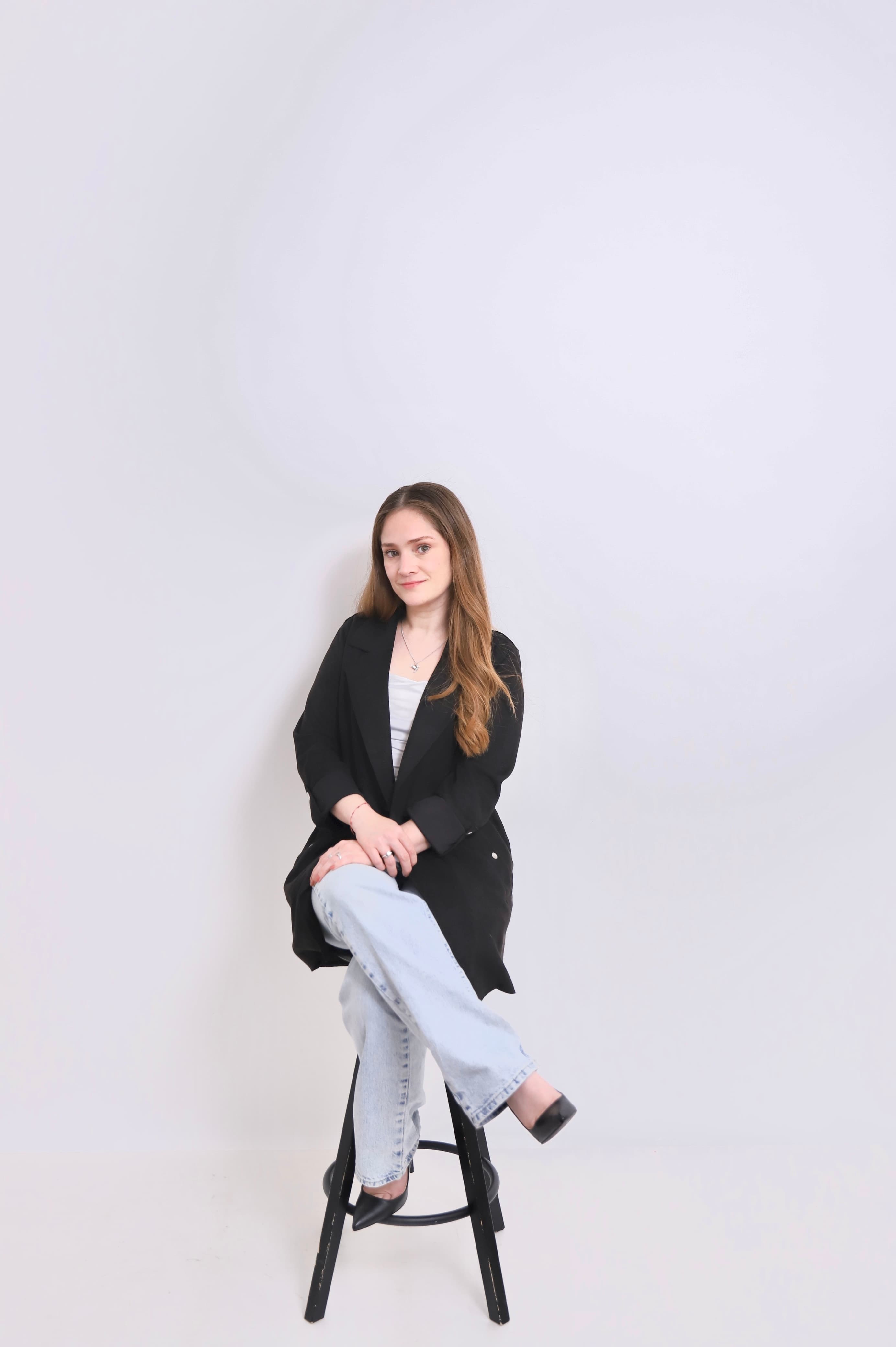 Woman with long brown hair wearing a black blazer and jeans sitting on a stool.