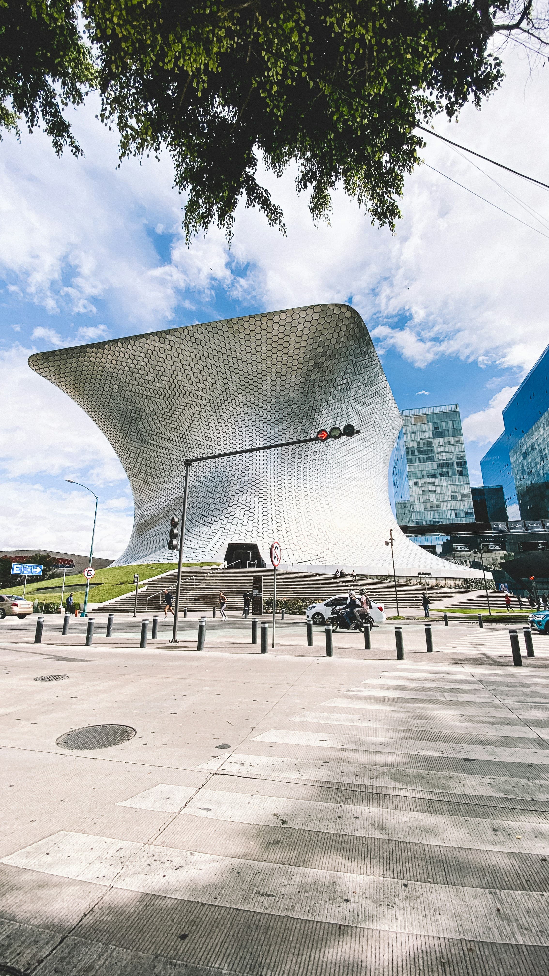 Curved, metallic Soumaya Museum with hexagonal tiles rises above a city street and crosswalk.